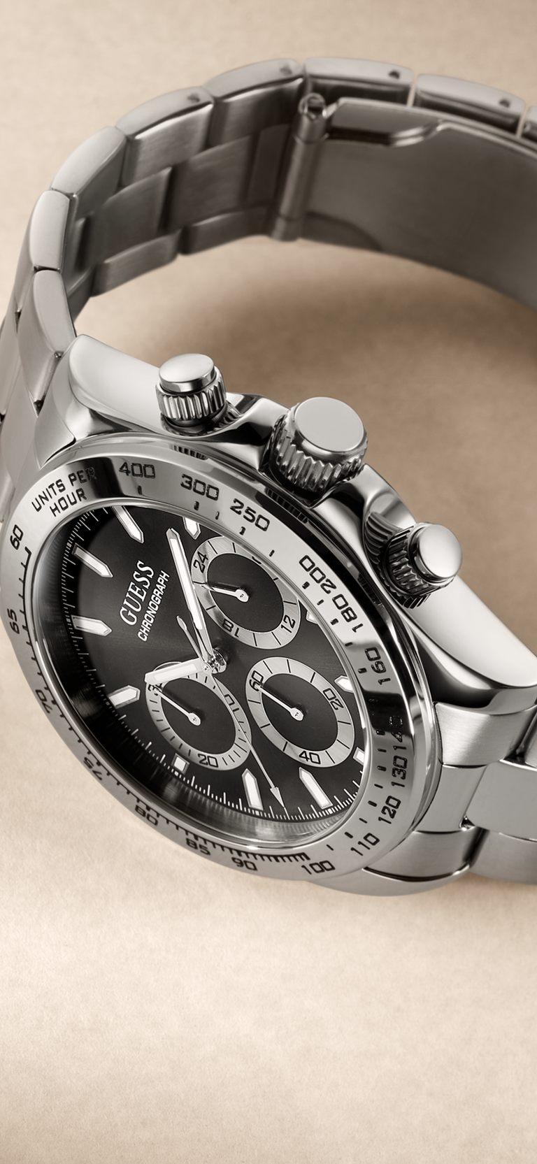 A Guess wrist watch with a silver stainless steel link bracelet, a black dial with three subdials, and a black tachymeter bezel, shown on a neutral beige background.