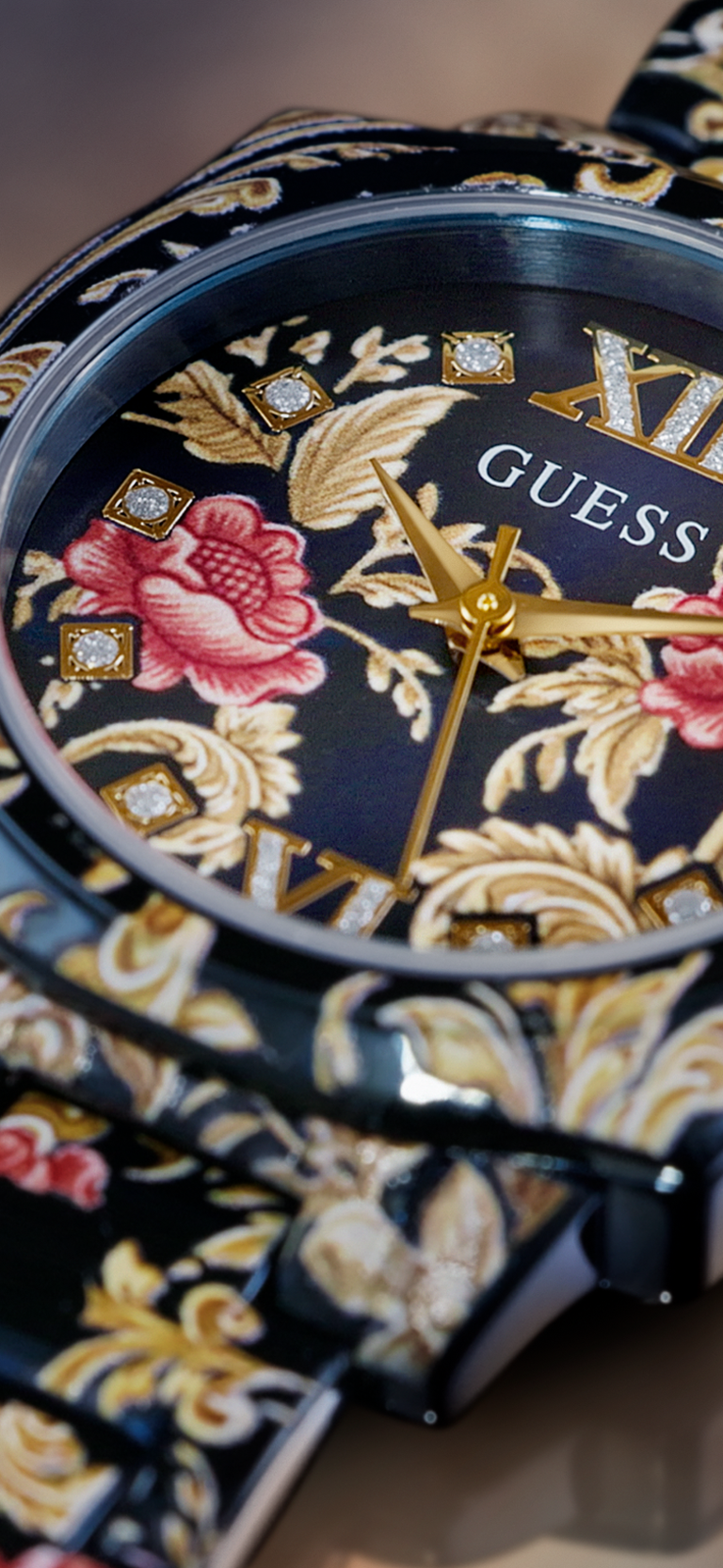 Close-up of a GUESS wristwatch with a black dial decorated with gold baroque-style floral motifs, pink rose details, and crystal accents, paired with a matching patterned strap.