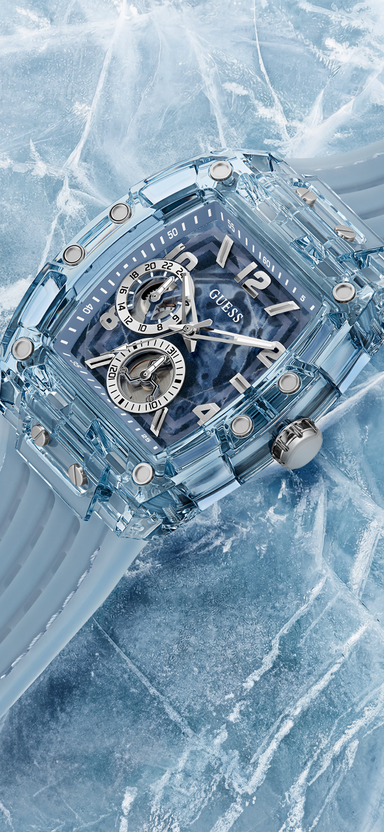 A GUESS chronograph watch with a transparent light blue resin case and strap, featuring a blue dial with silver-tone subdials and markers, set against a textured icy blue background.