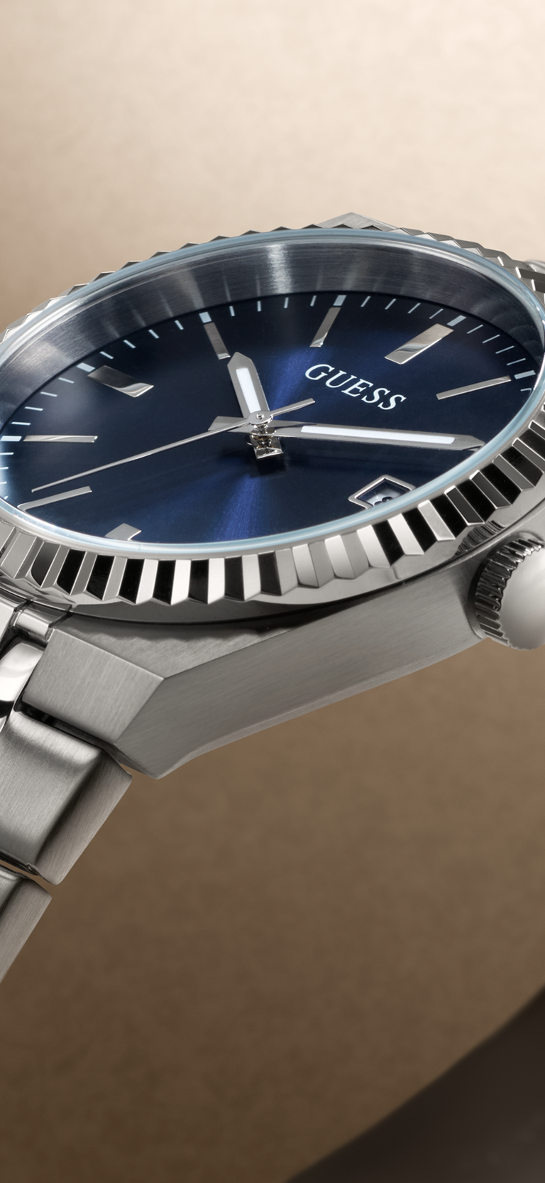 Close-up of a Guess wristwatch with a silver stainless steel case, fluted bezel, and blue dial, shown against a warm beige background.