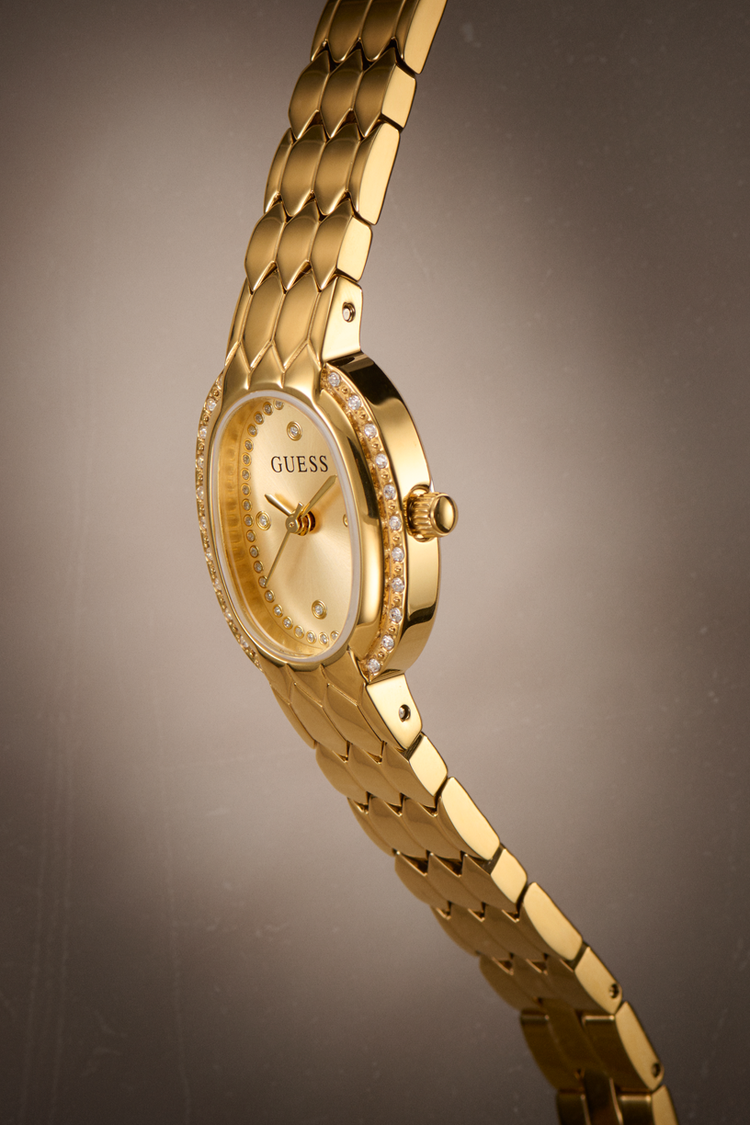 Gold-tone Guess women's watch with a metal link bracelet, diamond-accented bezel, and gold dial, shown on a dark gray gradient background.