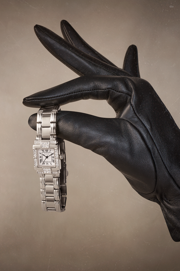 A silver-toned rectangular watch with a metal link bracelet is held between a black leather glove and fingers against a warm taupe background.
