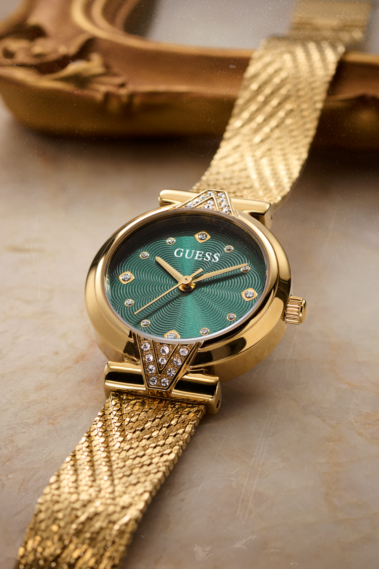 A gold-tone GUESS wristwatch with a textured green dial and a matching gold mesh strap, resting on a neutral stone surface with a blurred decorative gold object in the background.