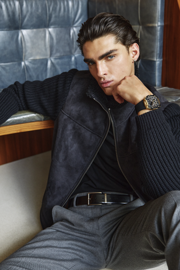 Man wearing a dark jacket and gray pants sitting against a textured blue wall.