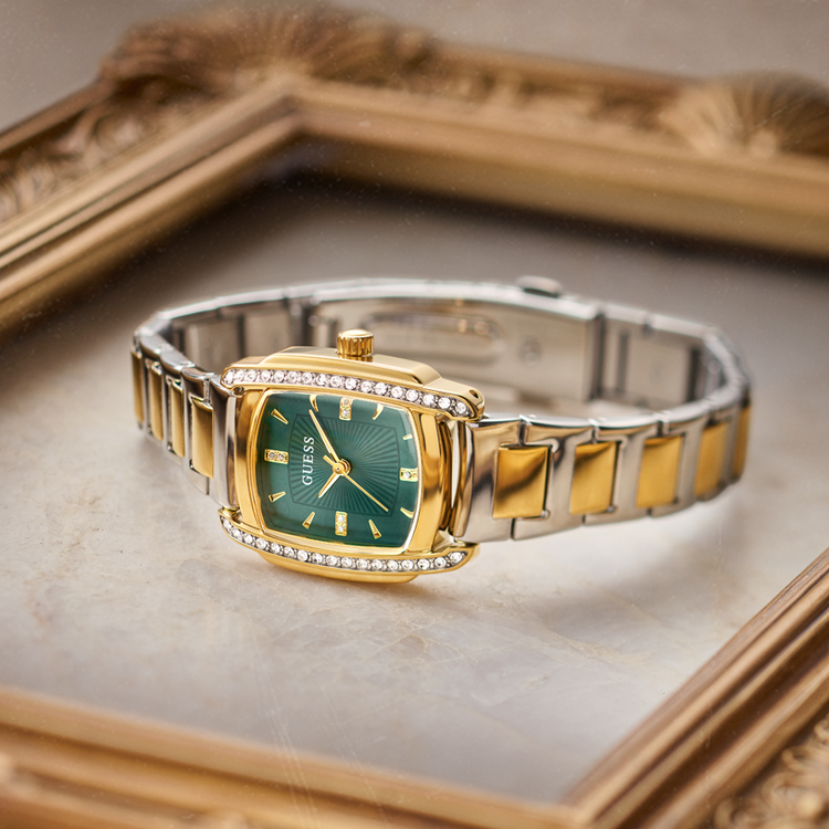 A Guess women's watch with a gold-tone and stainless steel link bracelet, featuring a green sunburst dial and crystal accents, displayed on a beige marble surface with an ornate gold picture frame in the background.