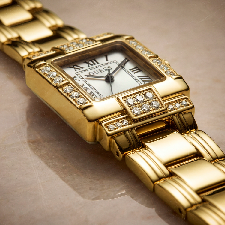 A gold-tone GUESS watch with a diamond-encrusted bezel and a white dial, shown in a close-up shot on a neutral beige surface.
