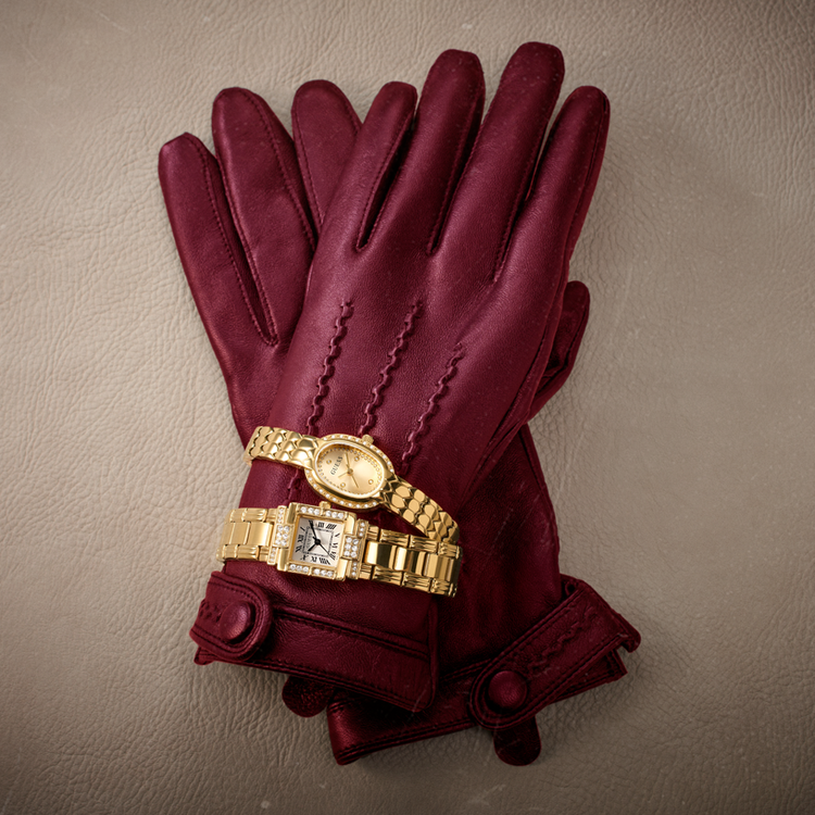 A pair of burgundy leather gloves with decorative stitching and cuff buttons, styled with two gold-tone metal wristwatches on a textured beige background.