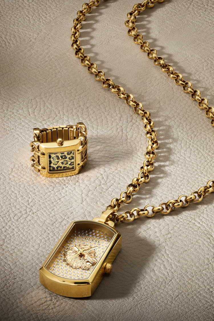 Gold-tone watch and matching ring set displayed on a textured beige surface. The watch features a rectangular dog-tag style case with a diamond-encrusted dial and a gold chain, while the ring has a similar rectangular face with a green and gold patterned bezel.