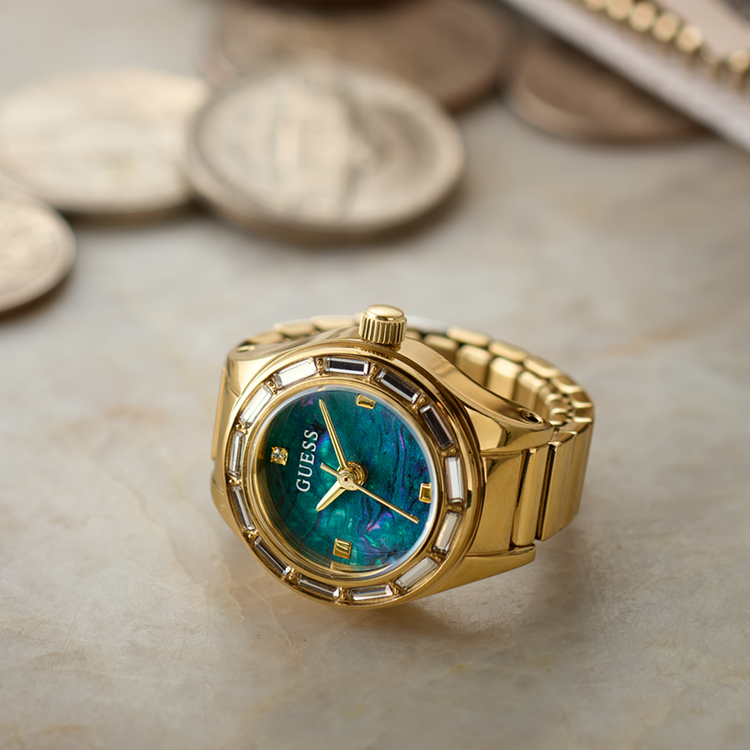 A gold-tone Guess watch with a blue-green abalone shell dial, resting on a light beige marble surface with blurred coins in the background.