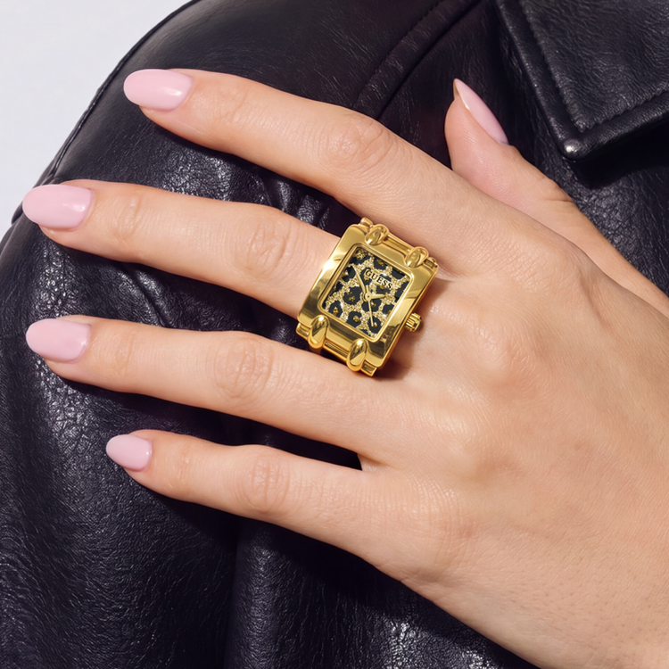 A gold-tone rectangular watch with a black patterned dial, worn on a hand with light pink manicured nails, resting against a black leather surface.