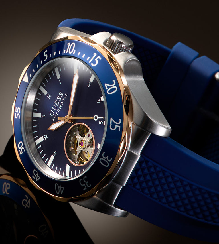 A close-up studio shot of a Guess Swatch automatic watch with a blue dial, blue bezel, blue strap, and silver-toned case with rose-gold accents.