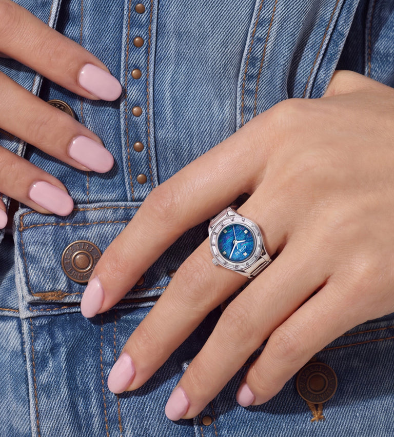 A close-up of a person's hand wearing a silver-toned ring with a blue watch face, resting against a blue denim jacket.