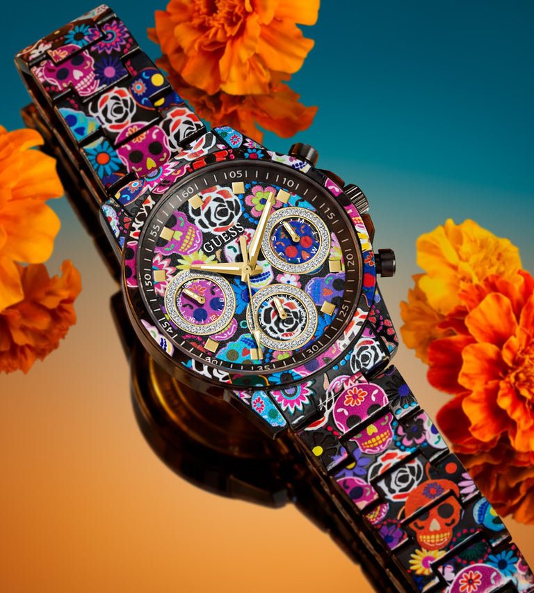 A GUESS chronograph watch with a colorful sugar skull and floral patterned dial and strap, displayed diagonally against a blue-to-orange gradient background with orange marigold flowers.