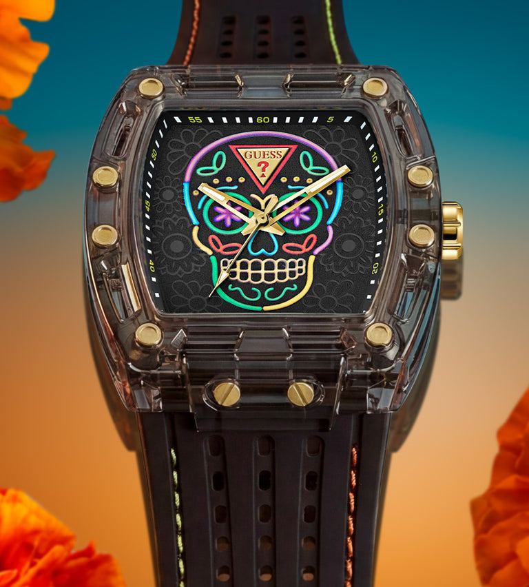 A Guess brand watch with a transparent dark gray case, gold-tone screws, and a black strap. The dial features a colorful sugar skull graphic with neon-style outlines in pink, blue, green, and yellow.
