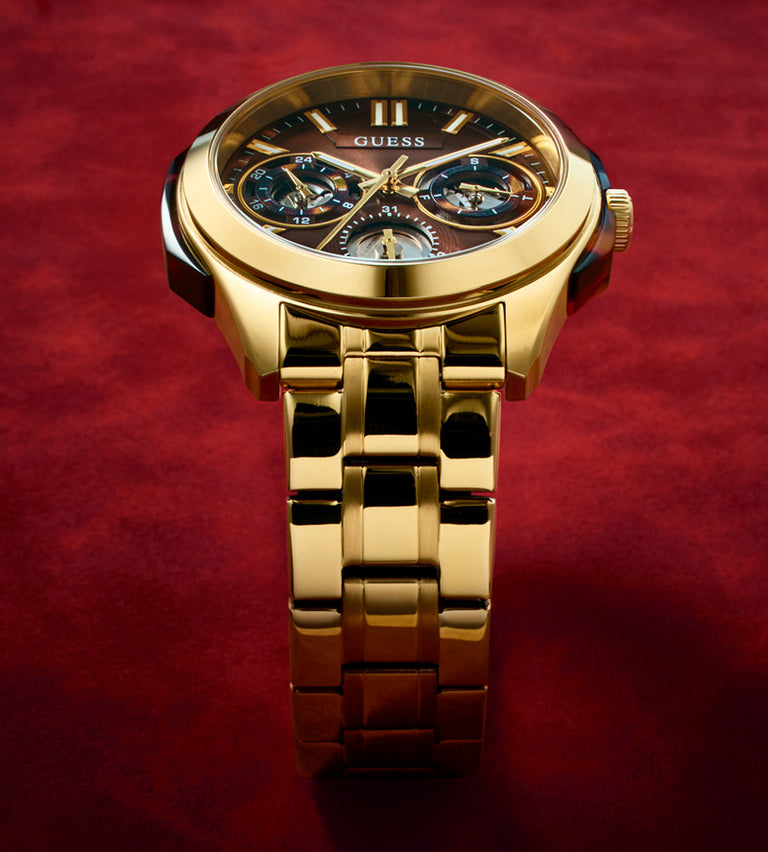Gold-tone Guess wristwatch with a multi-dial chronograph-style face and matching metal link bracelet, photographed on a rich red textured background.