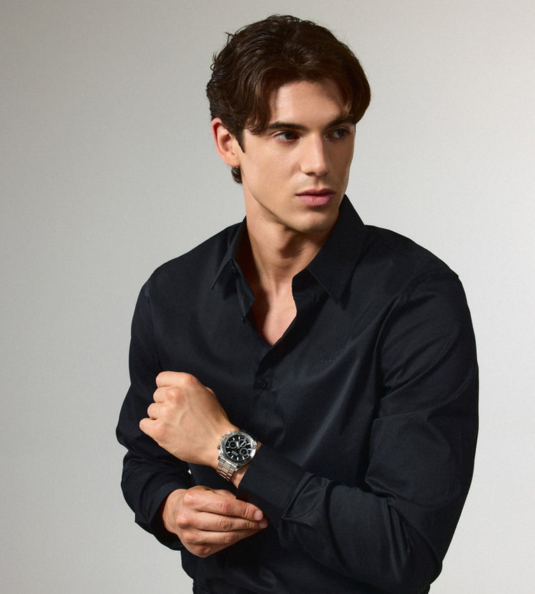 A man wearing a black long-sleeve button-down shirt and a silver-toned wristwatch with a black dial, posed against a light gray studio background.