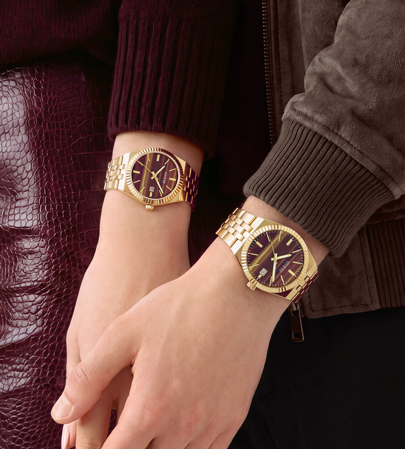 Two women's gold-tone metal bracelets with dark brown or black round dials, shown worn on two wrists. The image is a close-up lifestyle shot featuring the gold watch bracelets on models' wrists.