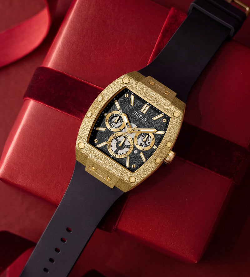 A luxury watch with a textured gold-tone case, black dial, and black strap, presented diagonally on top of a red gift box with a red ribbon.