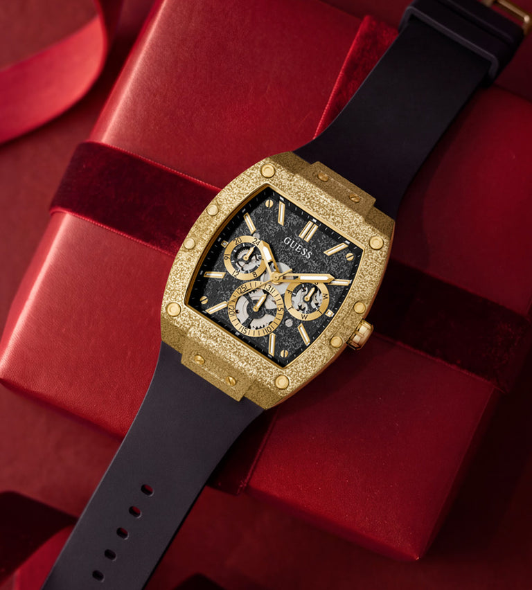 A luxury watch with a textured gold-tone case, black dial, and black strap, presented diagonally on top of a red gift box with a red ribbon.