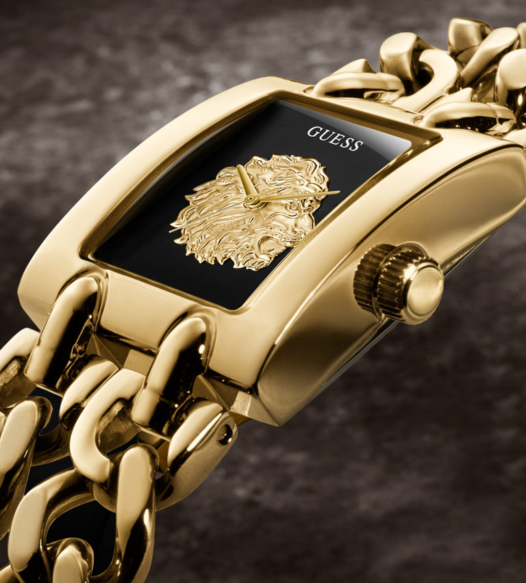 Gold-tone Guess watch with a rectangular black dial, raised gold medallion logo, and chunky gold chain bracelet on a dark textured background.