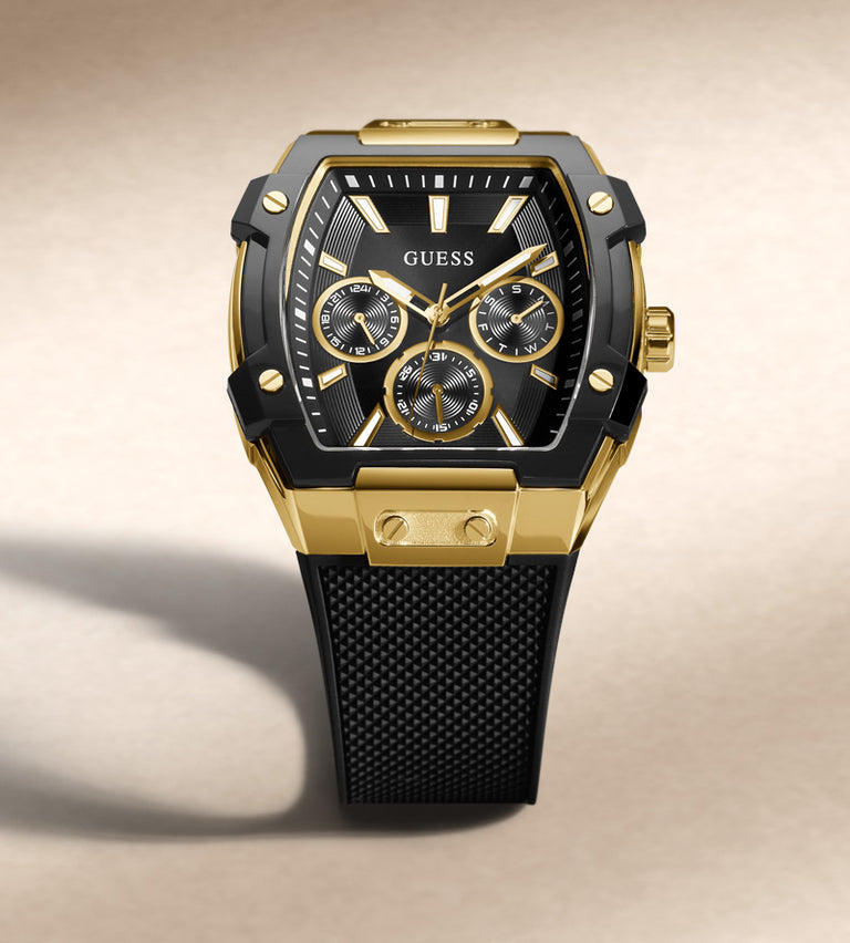 A GUESS men's watch featuring a black and gold tonneau-shaped case, a black dial with gold accents, and a textured black strap, set against a neutral beige background.