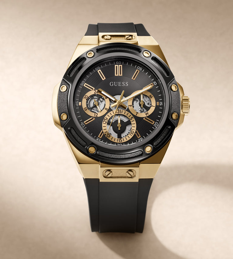 A luxury wristwatch by Guess with a gold-tone case, black bezel and dial, and a black rubber strap, shown on a warm beige background.