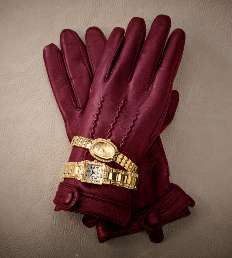A pair of burgundy leather gloves with decorative stitching and cuff details, styled with two gold-tone watches on a textured beige background.