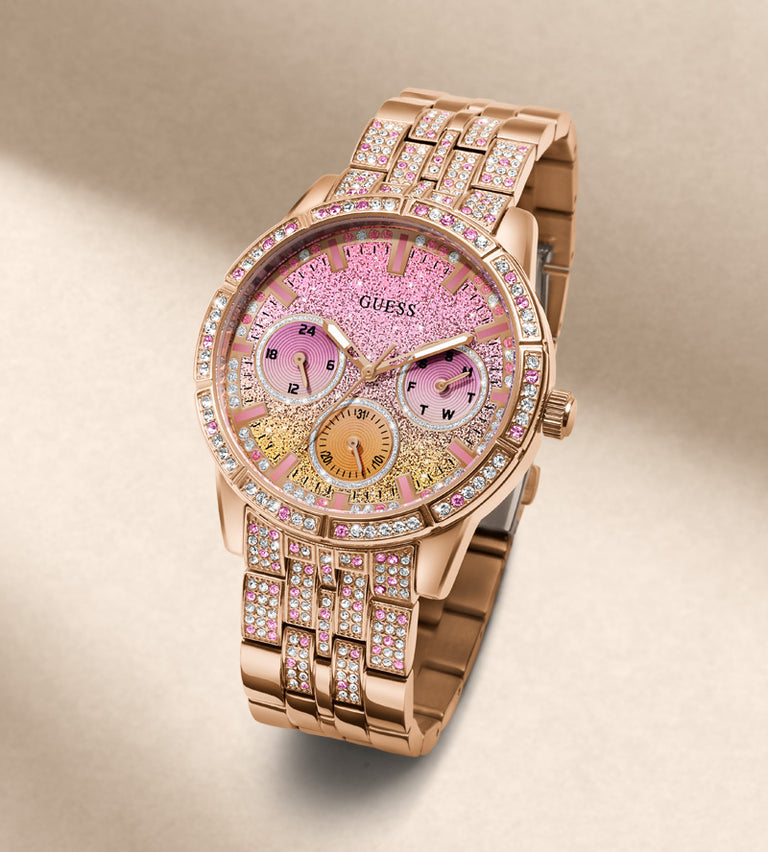 A luxury Guess watch featuring a rose gold metal bracelet and case, embellished with sparkling crystals. The dial is a glittery pink with gold-toned sub-dials and Roman numeral hour markers.