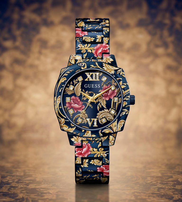 A Guess wristwatch with a dark blue floral dial and matching patterned strap, featuring gold-tone hands and Roman numerals, set against a textured brown background.