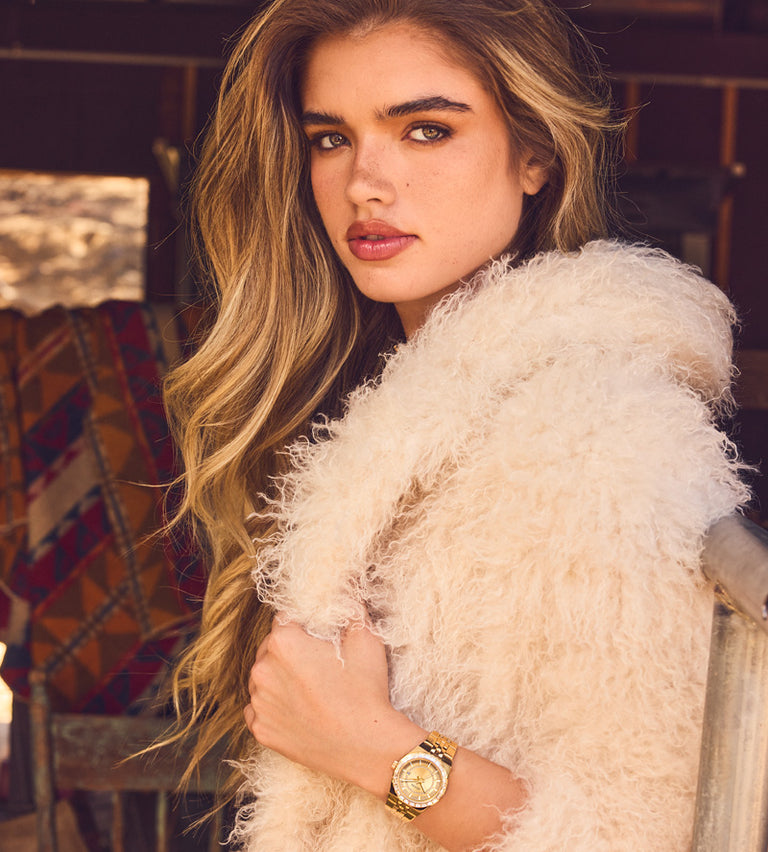 A woman with long wavy blonde hair wears a chunky cream faux fur wrap and a gold-tone watch with a gold dial, posed in a rustic indoor setting.