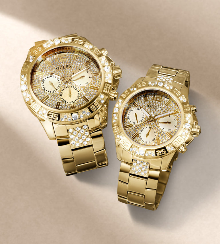 Two gold-tone Guess wristwatches with crystal-embellished bezels and bracelets, displayed on a warm beige background.