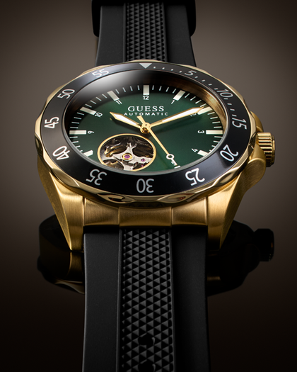 A luxury wristwatch by Guess with a gold-tone case, a dark green dial featuring a visible mechanical movement through a cutout, and a black bezel with white numerals, paired with a black textured rubber strap.