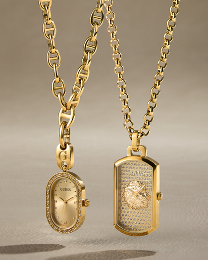 Two gold-tone Guess necklaces displayed against a neutral beige background. One necklace features an oval pendant with a diamond-encrusted bezel and a gold dial with 'GUESS' branding.