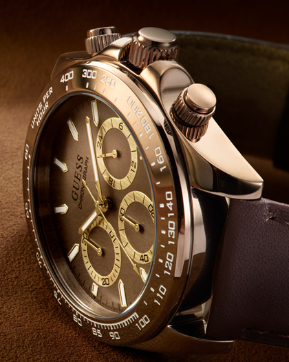 A close-up of a Guess wristwatch with a dark brown dial, gold-tone subdials and markers, a brushed silver-tone case, and a dark brown leather strap on a warm brown background.