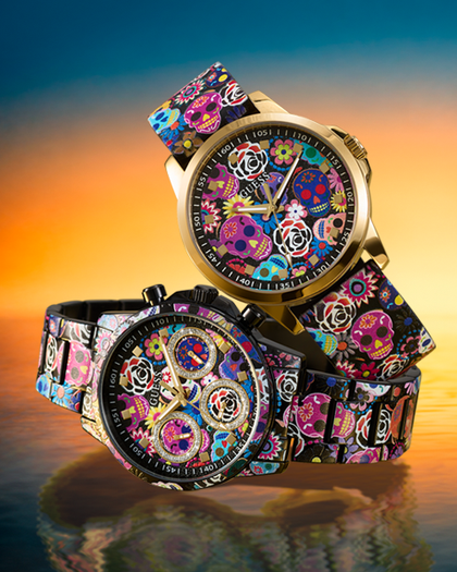 Two wristwatches with colorful floral and skull-patterned straps and gold-tone cases, displayed against a sunset-like gradient background with reflections below.