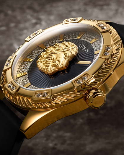Close-up of a gold-tone Guess wristwatch with a black strap, crystal-embellished bezel, and a sculpted gold lion head on the dial.