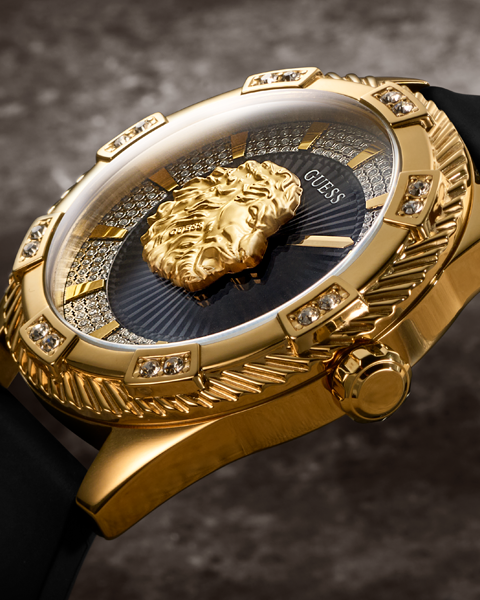 Close-up of a gold-tone Guess wristwatch with a black strap, crystal-embellished bezel, and a sculpted gold lion head on the dial.