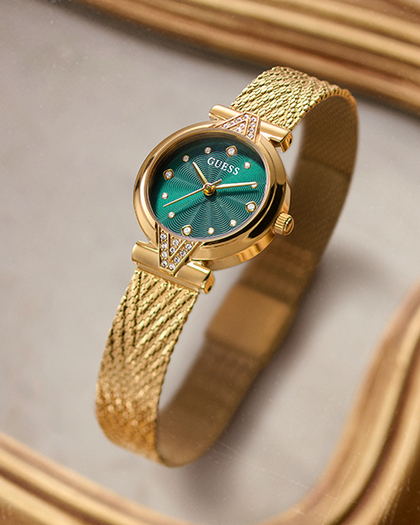 A gold-tone GUESS wristwatch with a textured mesh band and a green sunburst dial, resting on a reflective gold surface.