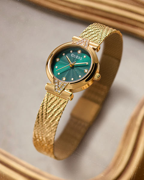 A gold-tone GUESS wristwatch with a textured mesh band and a green sunburst dial, resting on a reflective gold surface.