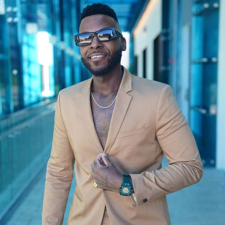 Man in a beige suit with sunglasses and a watch, standing in a modern building.