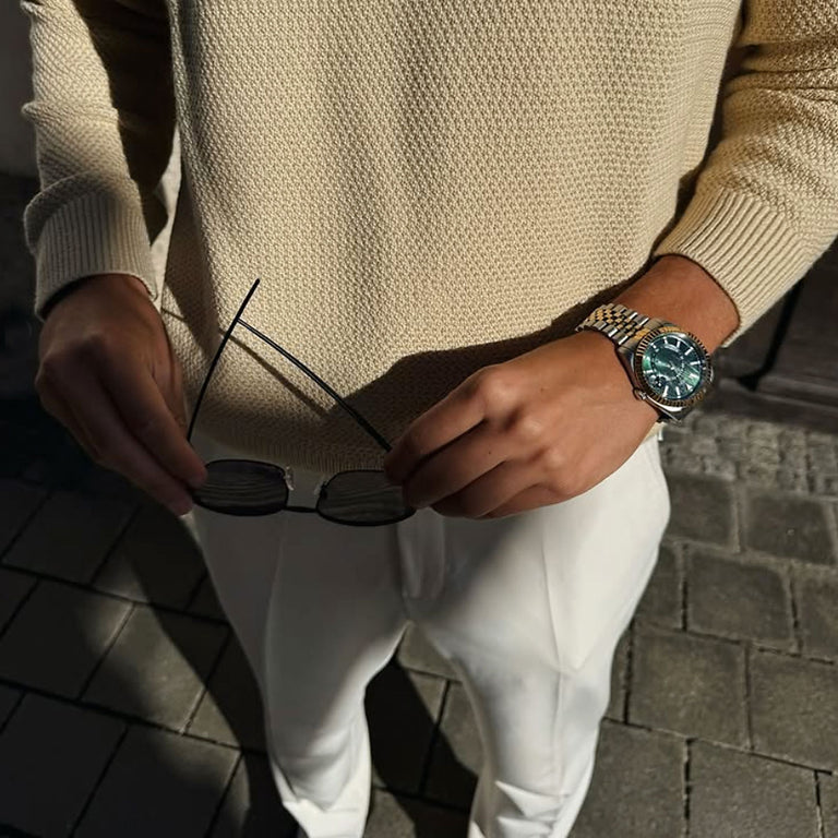 Person holding sunglasses with a watch on wrist, wearing a beige sweater and white pants.