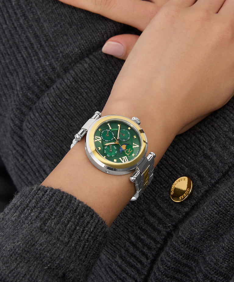 Close-up of a wrist wearing a Gc TopChic Large Size Metal watch with a green face and gold accents on a dark gray textured background.