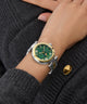 Close-up of a wrist wearing a Gc TopChic Large Size Metal watch with a green face and gold accents on a dark gray textured background.