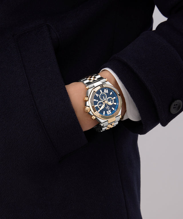 Close-up of a person wearing a Gc Atlas Chrono Metal watch with a blue face and gold accents, set against a dark coat.