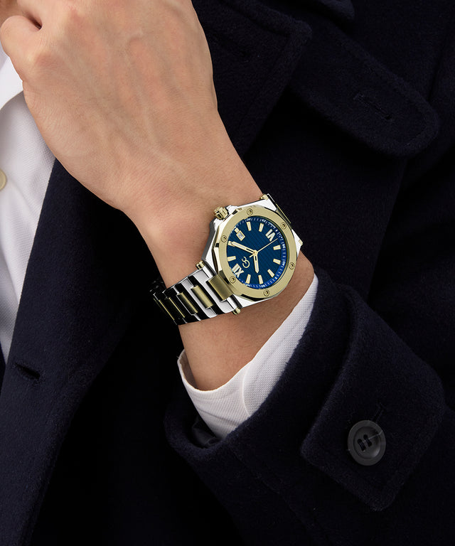 Person wearing a Gc Legacy Sleek Metal watch with a blue face and silver case, set against a dark coat and white shirt.