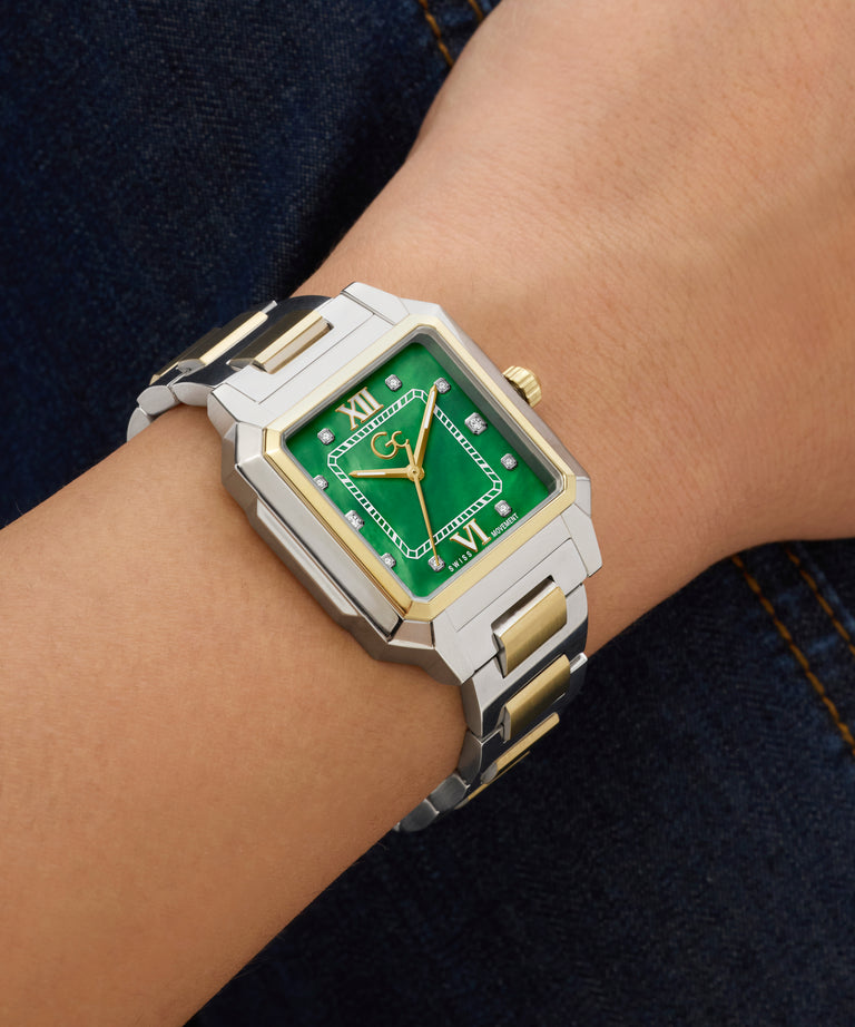 Wrist wearing a silver and gold Gc SquareCut Lady Mid Size Metal watch with a green face on a dark denim background