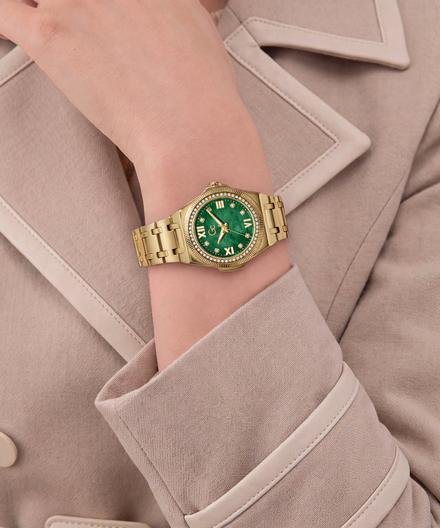 A gold-tone metal wristwatch with a green dial and bezel, worn on a model's wrist. The watch is styled with a beige trench coat.
