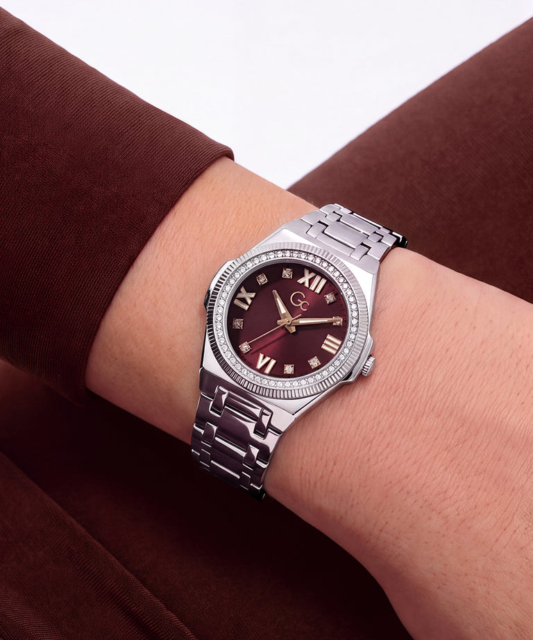 A silver-tone stainless steel watch with a dark burgundy dial and Roman numeral hour markers, shown on a person's wrist against a brown long-sleeve top.