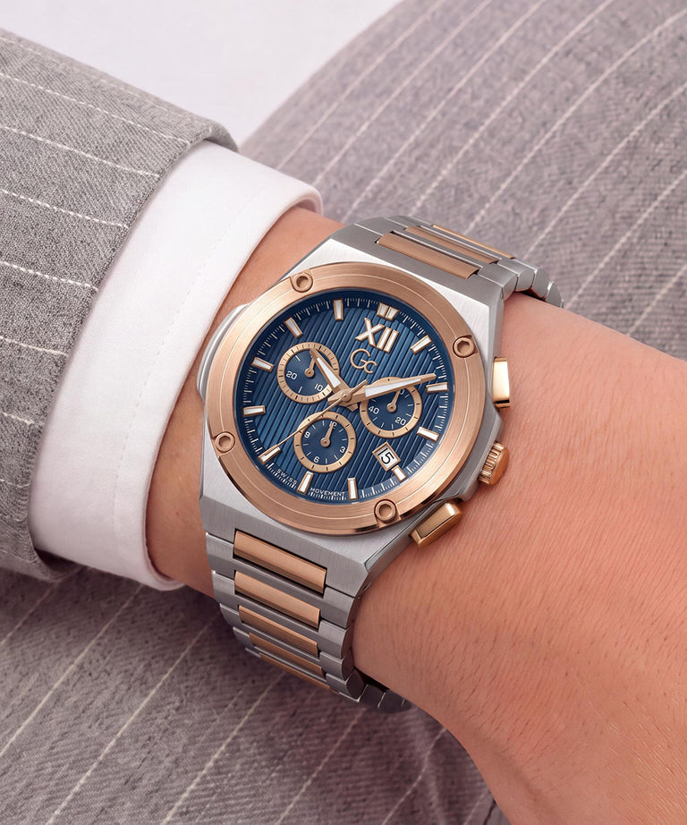 A two-tone stainless steel and rose gold chronograph watch with a blue sunburst dial, shown on a person's wrist wearing a white dress shirt and gray pinstripe suit jacket.