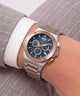 A two-tone stainless steel and rose gold chronograph watch with a blue sunburst dial, shown on a person's wrist wearing a white dress shirt and gray pinstripe suit jacket.